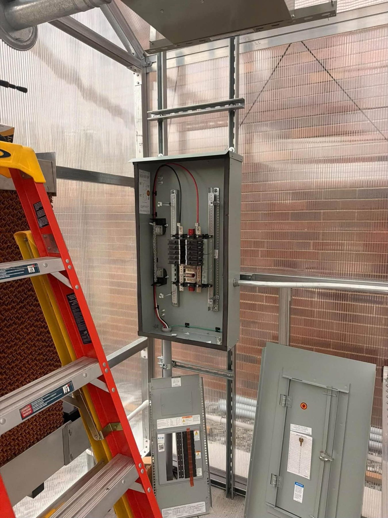 An open electrical panel being installed on metal strut, with an orange ladder beside it