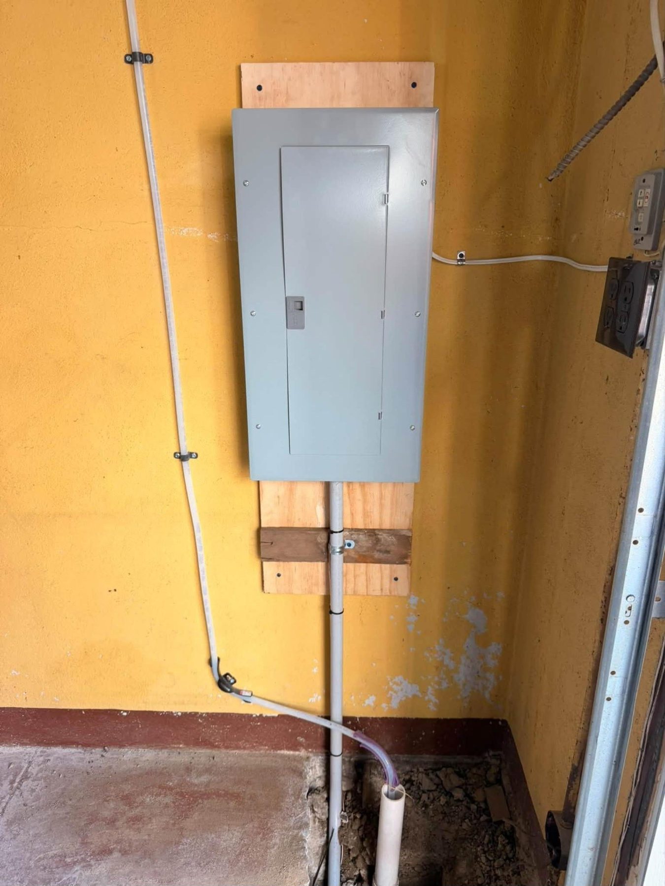 A new gray electrical panel mounted on plywood on a yellow garage wall with conduit running to the floor
