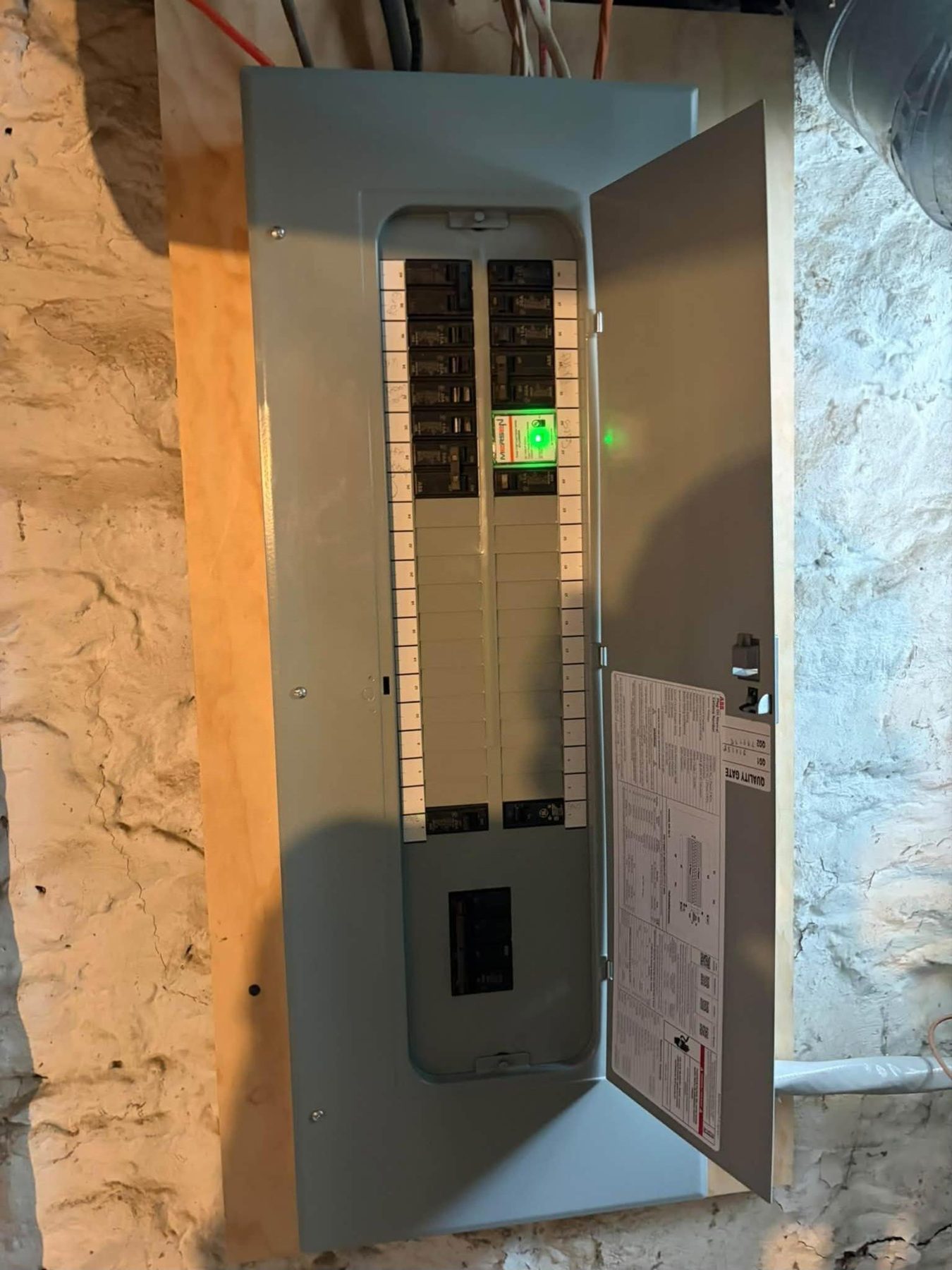 A new breaker panel with its door open and a surge protector glowing green, mounted on a stone basement wall